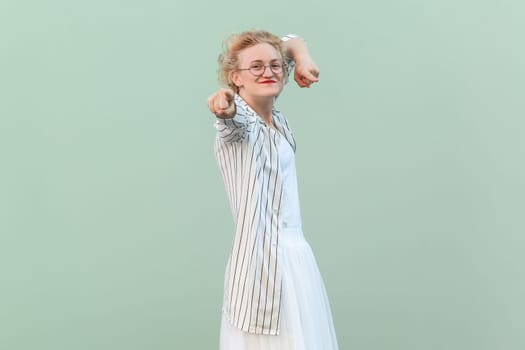 Portrait of joyful cheerful positive blonde woman wearing striped shirt and skirt, pointing index finger to camera, choosing you. Indoor studio shot isolated on light green background.