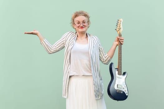 Portrait of uncertain blonde woman in shirt, skirt, and striped blouse with eyeglasses holding electric guitar, spreads hand, looks confused. Indoor studio shot isolated on light green background.