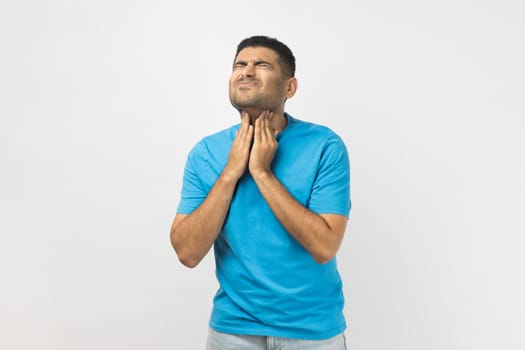 Portrait of sick sad unshaven man wearing blue T- shirt standing touching his painful neck, having sore throat, frowning face, being very sick. Indoor studio shot isolated on gray background.
