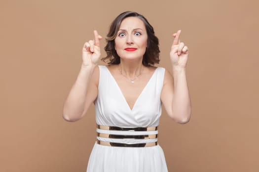 Portrait of pleasant looking hopeful middle aged woman with wavy hair crosses fingers and has great hope for better, wearing white dress. Indoor studio shot isolated on light brown background.