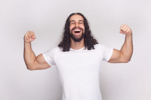 Portrait of happy cheerful strong bodybuilder man in white t-shirt with long wavy hair and beard, standing showing his biceps and triceps. Indoor studio shot isolated on gray background.