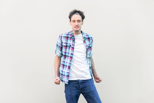 Portrait of angry aggressive handsome man standing with clenched fists, arguing with somebody, wearing blue checkered shirt and headband. Indoor studio shot isolated on gray background.