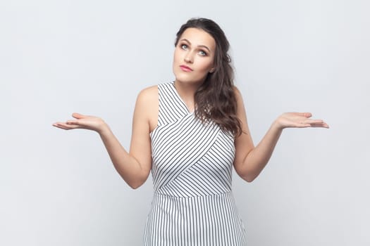 Portrait of brunette woman spreads palms, shrugs shoulders with perplexed expression, being indecisive, has no idea what happened, wearing striped dress. Indoor studio shot isolated on gray background