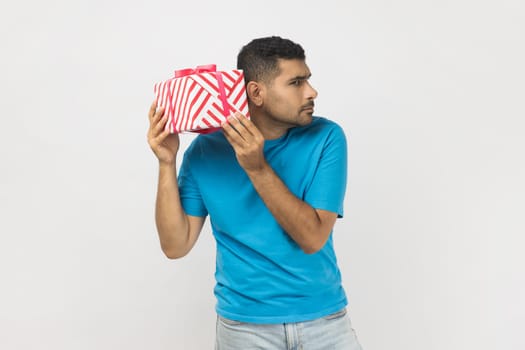 Portrait of curious unshaven man wearing blue T- shirt standing with interested facial expression, shaking present box, thinking what inside. Indoor studio shot isolated on gray background.