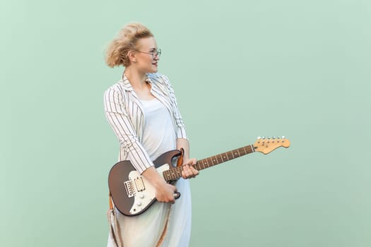 Portrait of beautiful positive blonde woman in white shirt, skirt, and striped blouse with eyeglasses, holding guitar and looking away. Indoor studio shot isolated on light green background.