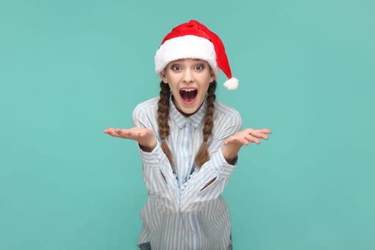 Portrait of excited amazed teenager girl with braids wearing striped shirt and Santa Claus hat, looking at camera with open mouth, spread hands. Indoor studio shot isolated on green background.