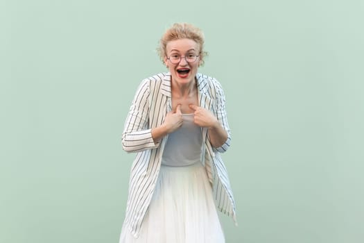 Portrait of surprised astonished excited blonde woman wearing striped shirt and skirt, pointing at herself, being amazed. Indoor studio shot isolated on light green background.