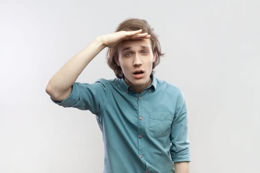 Portrait of concentrated young man looking far away at distance with hand over head, attentively searching for bright future, wearing blue shirt. Indoor studio shot isolated on gray background.