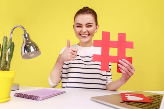 Smiling woman blogger in good mood, showing red hashtag and thumb up like gesture, taking selfie POV, sitting at workplace. Indoor studio studio shot isolated on yellow background.