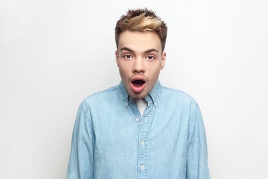 Portrait of amazed surprised man wearing denim shirt looking at camera with big eyes and open mouth, sees something shocked and astonished. Indoor studio shot isolated on gray background.