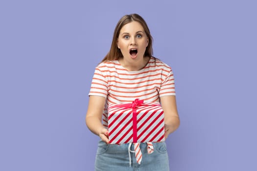 Portrait of astonished excited blond woman wearing striped T-shirt giving gift box with amazed facial expression, looking at camera with big eyes. Indoor studio shot isolated on purple background.