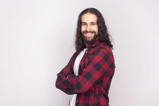 Side view portrait of smiling happy bearded man with long curly hair in checkered red shirt standing with crossed arms, expressing confidence. Indoor studio shot isolated on gray background.