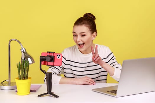 Satisfied woman blogger in striped shirt recording video for followers, looking at phone camera, talking with followers, showing v sign. Indoor studio studio shot isolated on yellow background.