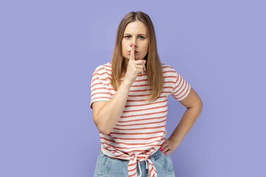 Shh, be quiet. Portrait of serious woman wearing striped T-shirt showing silence gesture with finger on her mouth, asking to stay quiet, keep secret. Indoor studio shot isolated on purple background.