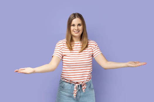 Friendly positive optimistic blond woman wearing striped T-shirt standing with raised arms, looking with toothy smile, offering to take for free. Indoor studio shot isolated on purple background.