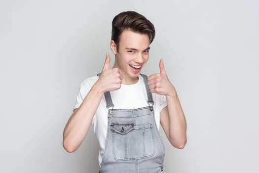 Nice joke. Portrait of young brunette man standing keeps thumb raised, being in good mood, shows his agreement, wearing denim overalls. Indoor studio shot isolated on gray background.
