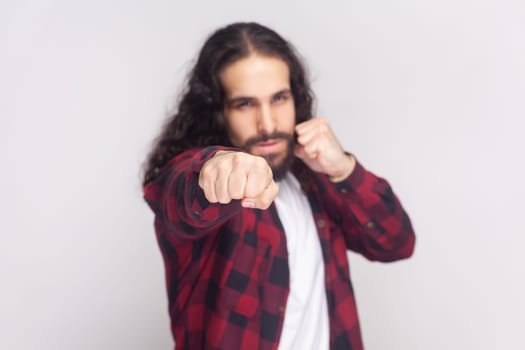 Aggressive bearded man with long curly hair in checkered red shirt keeping his fists ready to fight and defense himself against injustice or violence. Indoor studio shot isolated on gray background.