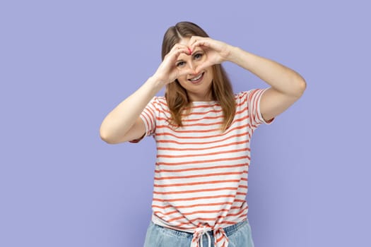 Portrait of blond woman wearing striped T-shirt making heart shape with hands in front of her eyes, expressing love, friendship, care and romance. Indoor studio shot isolated on purple background.