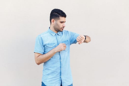 Portrait of serious concentrated man wearing denim shirt standing looking at his wristwatch, being busy, checking time, timing. Indoor studio shot isolated on gray background.
