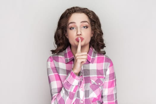 Pleasant looking delightful woman with curly hair keeps finger on lips, asks not tell secret information or keep silence, wearing pink checkered shirt. Indoor studio shot isolated on gray background.