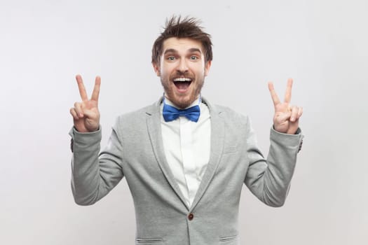 Excited satisfied delighted bearded man showing v sign, victory gesture, looking at camera with amazed expression, wearing grey suit and blue bow tie. Indoor studio shot isolated on gray background.