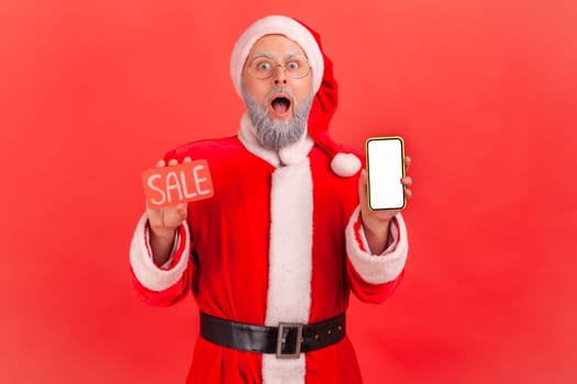 Portrait of shocked elderly man with gray beard wearing santa claus costume holding smart phone with empty screen and sale card, huge discounts. Indoor studio shot isolated on red background.