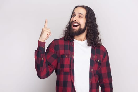 Portrait of amazed handsome bearded man with long curly hair in checkered red shirt raises fore finger as gets some brilliant idea. Indoor studio shot isolated on gray background.