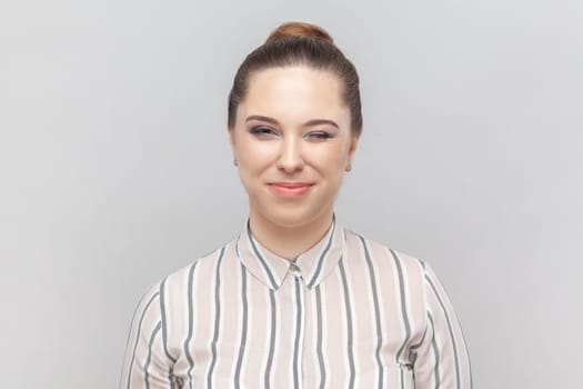 Portrait of beautiful playful woman wearing striped shirt standing looking at camera and winking, flirting with her boyfriend, smiling happily. Indoor studio shot isolated on gray background.