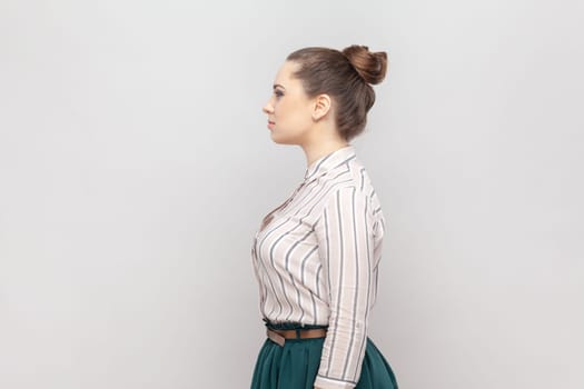 Side view portrait of serious strict woman with bun hairstyle wearing striped shirt and green skirt standing looking ahead, having bossy expression. Indoor studio shot isolated on gray background.