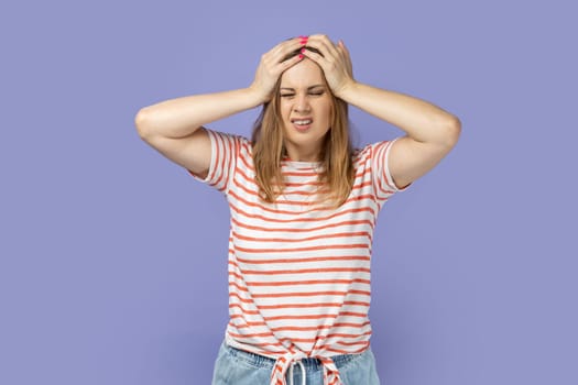 Portrait of unhealthy sick young adult blond woman wearing striped T-shirt massaging temples, suffering from headache, migraine. Indoor studio shot isolated on purple background.