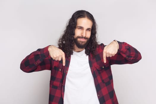 Portrait of bossy anger bearded man with long curly hair in checkered red shirt pointing down and looking arrogant, demanding to do immediately. Indoor studio shot isolated on gray background.