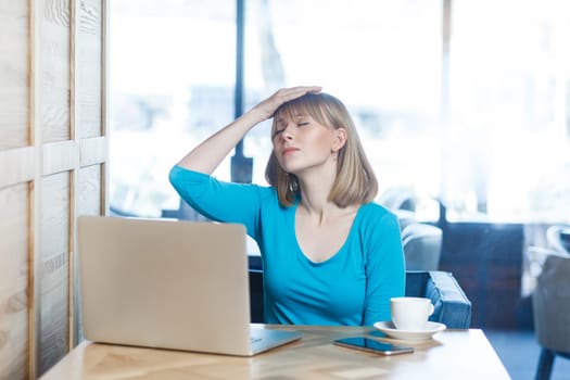 Portrait of sad upset despair young woman with blonde hair in blue shirt working on laptop, making facepalm gesture, has problems with work. Indoor shot in cafe with big window on background.