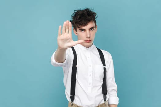 Portrait of strict serious young man wearing white shirt and suspender showing stop prohibition gesture with his palm, looking at camera. Indoor studio shot isolated on blue background.