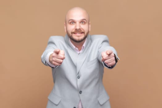 Portrait of cheerful bald bearded man pointing to you, has joyful expression, looking at camera with toothy smile, wearing gray jacket. Indoor studio shot isolated on brown background.