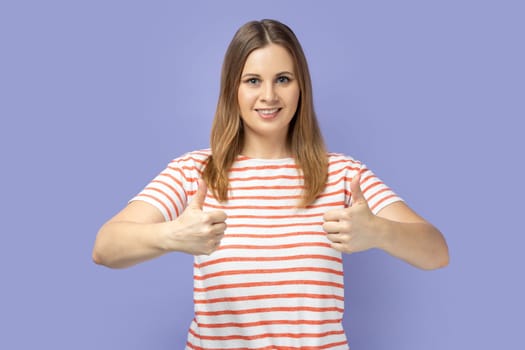 Portrait of blond woman wearing striped T-shirt looking at camera with toothy smile and showing thumbs up, approval sign, satisfied with service. Indoor studio shot isolated on purple background.