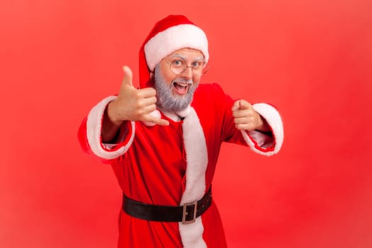 Elderly man with gray beard wearing santa claus costume holding fingers near ear showing telephone gesture, pointing at camera, waiting for your call. Indoor studio shot isolated on red background.