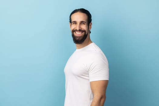 Side view of bearded handsome man wearing white T-shirt standing looking at camera with satisfied face and smiling, expressing happiness. Indoor studio shot isolated on blue background.