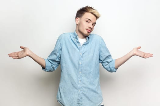Portrait of doubtful puzzled attractive man wearing denim shirt standing with spread hands, looking at camera and shrugging shoulders. Indoor studio shot isolated on gray background.