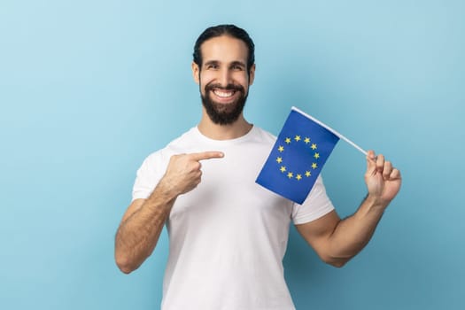 Portrait of man with beard wearing white T-shirt smiling broadly and pointing flag of European Union, symbol of Europe, EU association and community. Indoor studio shot isolated on blue background.