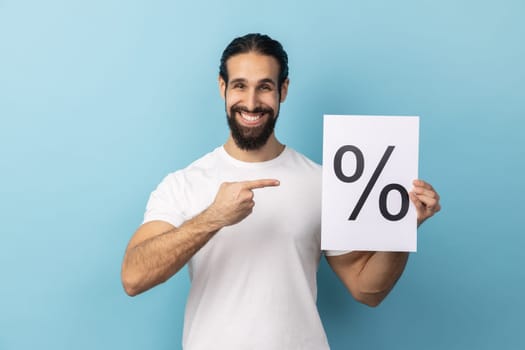 Portrait of attractive optimistic man with beard wearing white T-shirt holding and pointing paper with percent sign inscription, looking at camera. Indoor studio shot isolated on blue background.