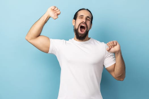 Portrait of sleepy handsome man with beard wearing white T-shirt standing and yawning with closed eyes and raised arms, waking up early. Indoor studio shot isolated on blue background.
