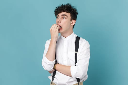 Portrait of panicking man wearing white shirt and suspender, biting nails, being nervous terrified, feeling frightened of challenge to start business. Indoor studio shot isolated on blue background.