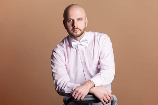 Portrait of handsome attractive bald bearded man looking at camera with serious concentrated facial expression, wearing light pink shirt and bow tie. Indoor studio shot isolated on brown background.