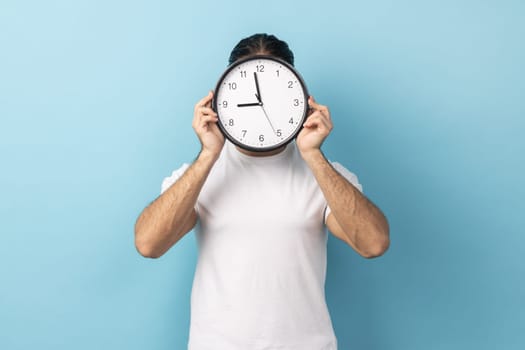 Portrait of anonymous man wearing white T-shirt holding wall clock hiding her face, time management, schedule and meeting appointment. Indoor studio shot isolated on blue background.