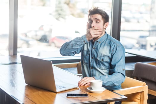 Portrait of handsome shocked scared young man freelancer in blue jeans shirt working on laptop, looking at screen, covering mouth with palm. Indoor shot near big window, cafe background.