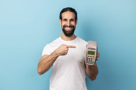 Portrait of joyful man with beard wearing white T-shirt pointing finger at pos terminal, suggesting you to use contactless payments. Indoor studio shot isolated on blue background.