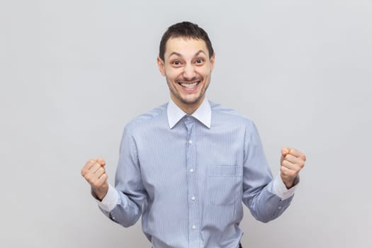 Portrait of extremely happy joyful cheerful man standing with clenched fists, rejoices his success, celebrating victory, wearing light blue shirt. Indoor studio shot isolated on gray background.