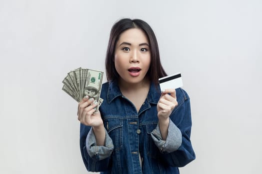 Portrait of shocked surprised brunette woman in blue denim jacket standing looking at camera with open mouth, holding dollar banknotes and credit card. Indoor studio shot isolated on gray background.