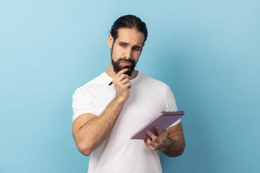 Portrait of thoughtful man with beard wearing white T-shirt holding paper notebook, having thoughtful facial expression, planning. Indoor studio shot isolated on blue background.