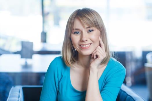 Portrait of smiling happy satisfied young woman with blonde hair in blue shirt sitting alone, looking at camera with toothy smile, being in good mood. Indoor shot in cafe with big window on background